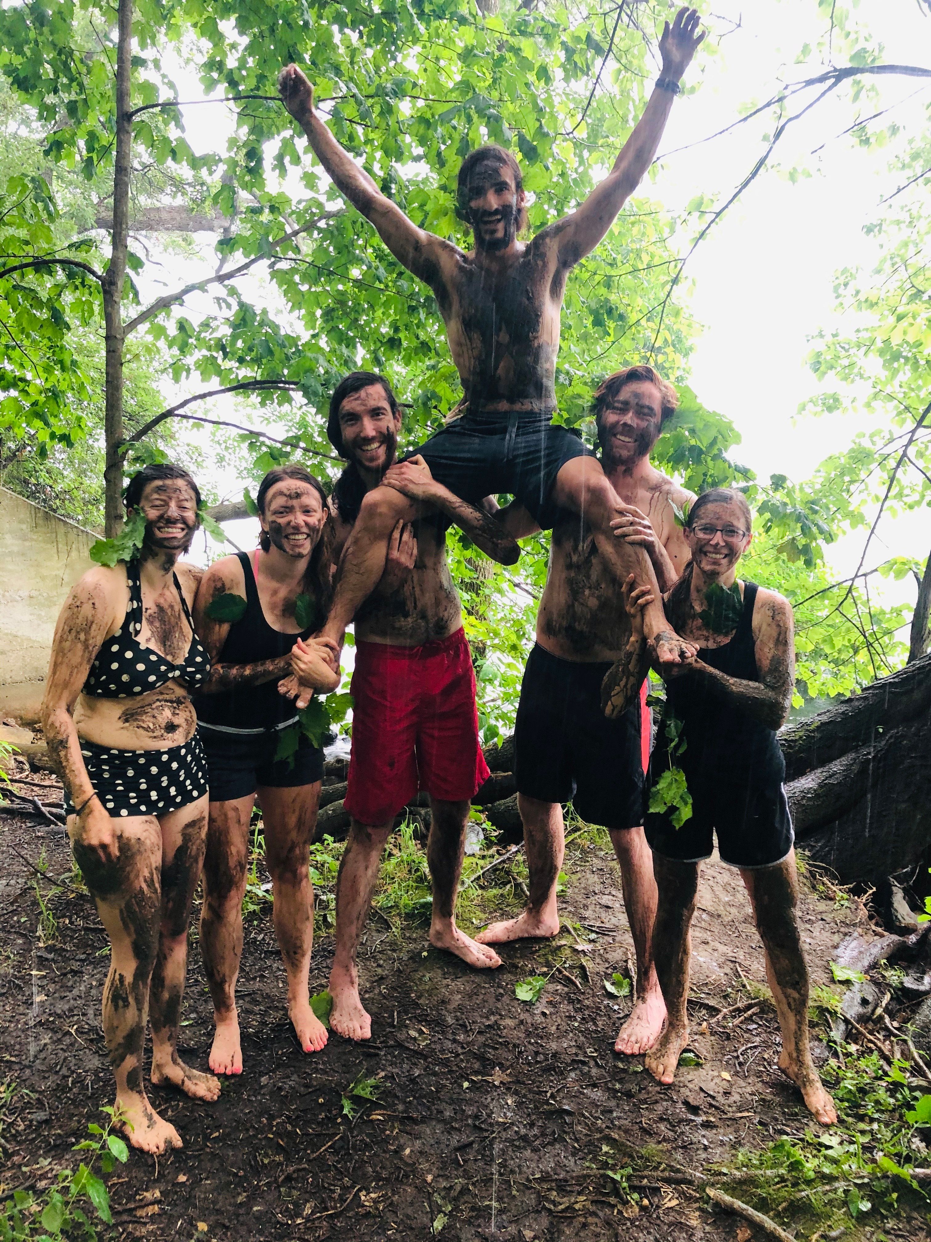 Muddy Summiteers in swimsuits stand triumphant to celebrate the summer solstice. One member is held help by the others, arms outstretched celebrating. (Circa summer 2020)